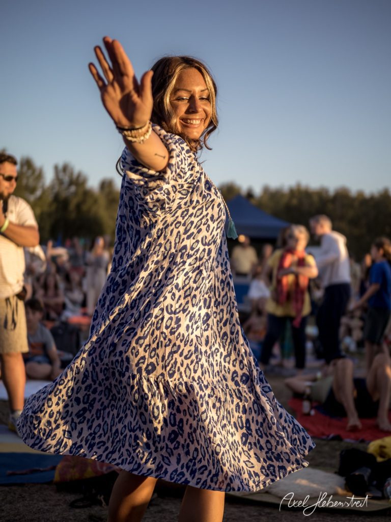 Eine fröhliche Frau in einem Leopardenkleid tanzt bei Sonnenuntergang auf einem Festivalgelände. – © Axel Hebenstreit