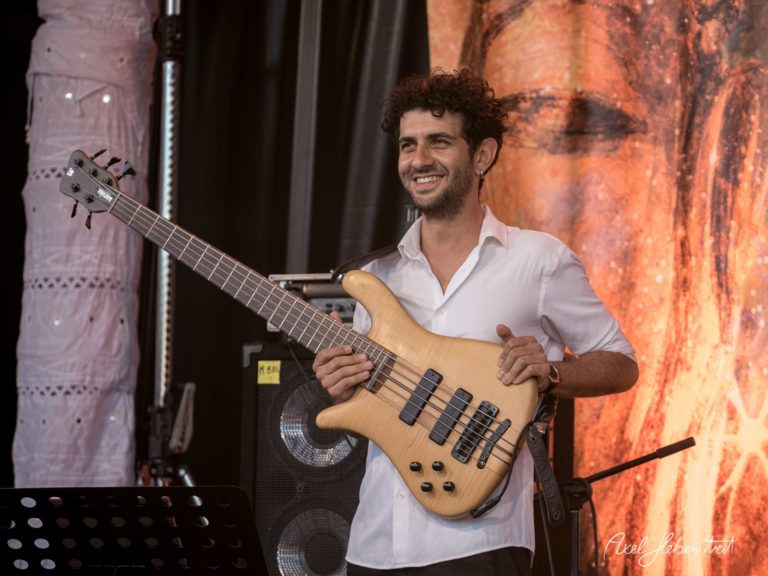 Musiker mit einem E-Bass lächelt auf einer Festivalbühne und strahlt Freude aus – © Axel Hebenstreit