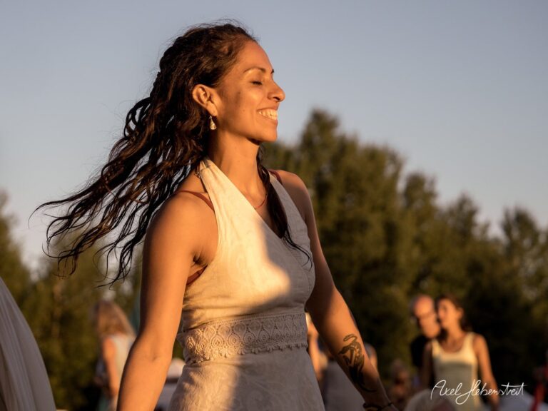 Eine lächelnde Frau tanzt bei Sonnenuntergang in einem Festivalambiente – © Axel Hebenstreit
