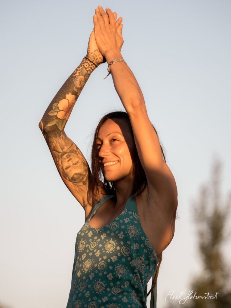 Eine lächelnde Frau mit artistischen Tattoos, die ihre Arme zum Himmel hebt, vor einem klaren Himmel – © Axel Hebenstreit