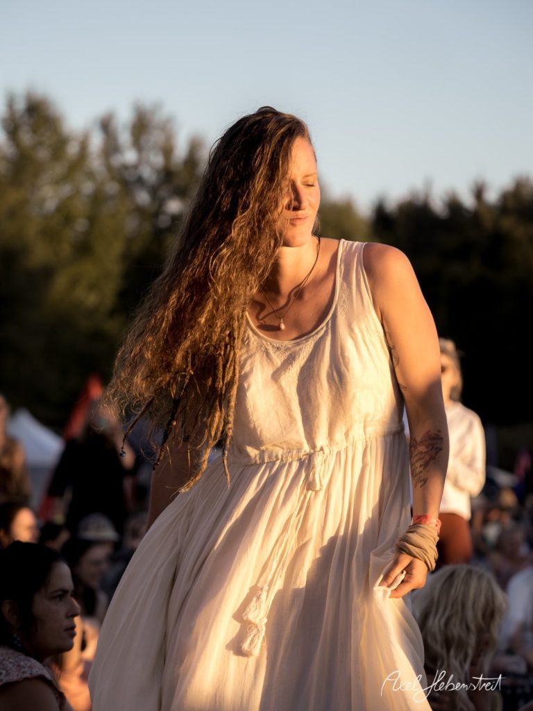 Frau in weißem Kleid tanzt im Sonnenlicht bei einem Festival in der Natur – © Axel Hebenstreit