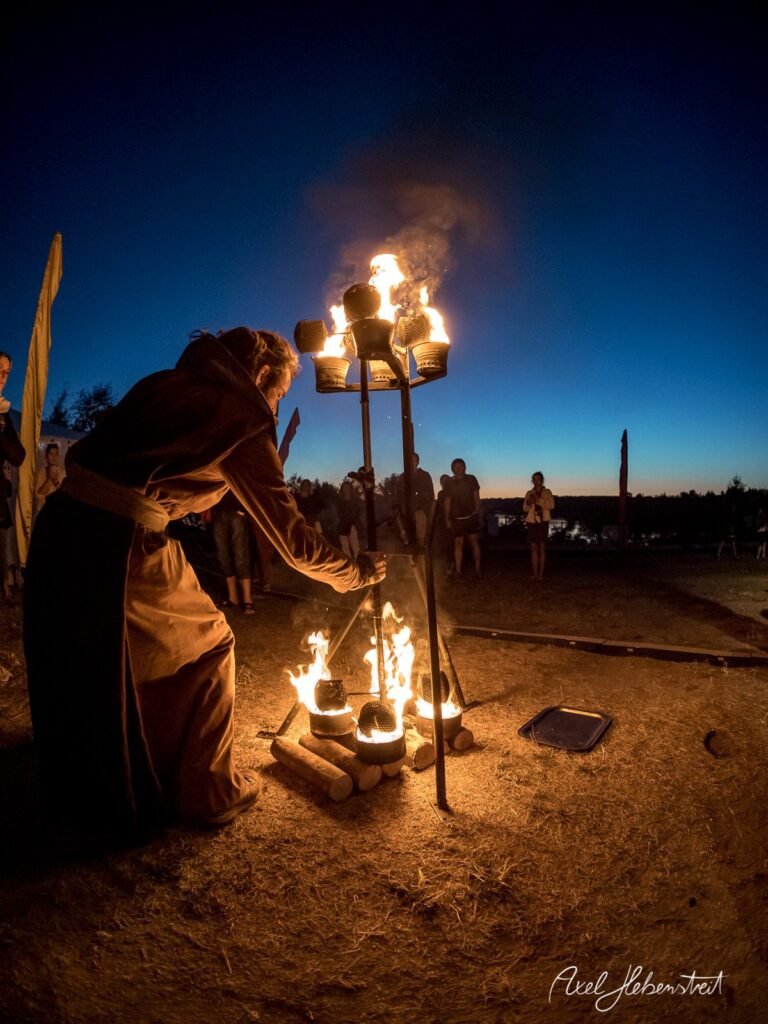 Ein Feuerzeremoniell in der Dämmerung mit einer Person, die ein brennendes Gefäß an zündet – © Axel Hebenstreit