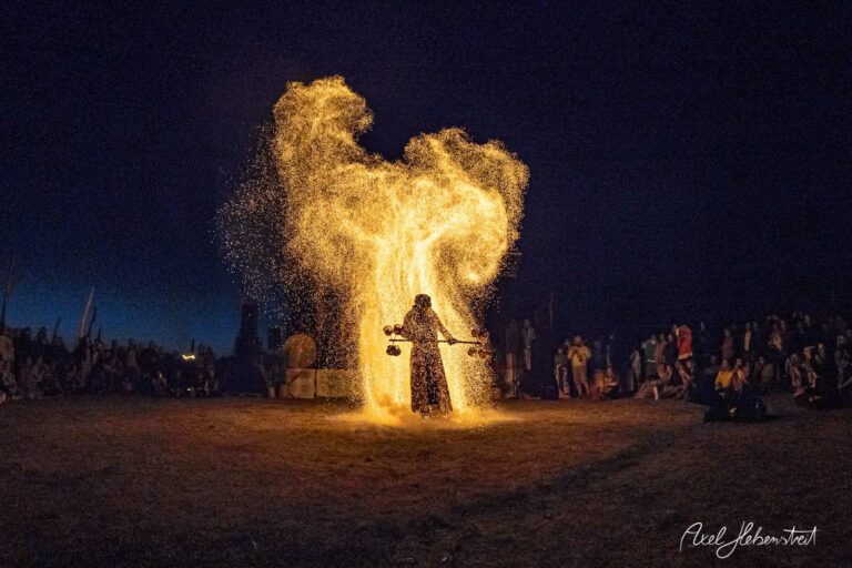 Eine Person in schwarzer Kleidung entfesselt funkelnde Funken in einer nächtlichen Festivalatmosphäre – © Axel Hebenstreit