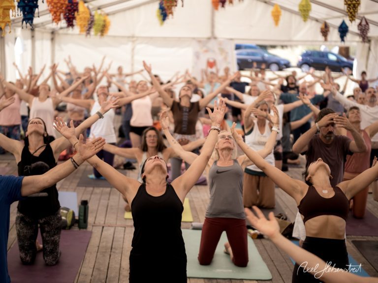 Eine Vielzahl von Teilnehmern in einem Zelt praktiziert energisch Yoga, mit erhobenen Armen in einer spirituellen Atmosphäre – © Axel Hebenstreit