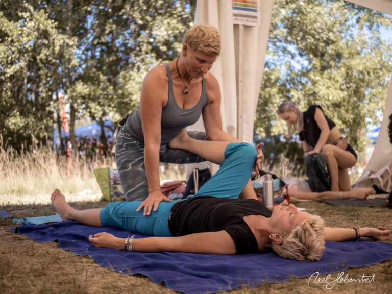 Zwei Frauen praktizieren Yoga bei einem Festival im Freien, wobei eine die andere dehnt – © Axel Hebenstreit.