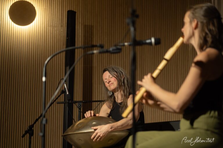 Zwei Musikerinnen spielen Handpan und Flöte in einem stimmungsvoll beleuchteten Studio mit Holzwand.