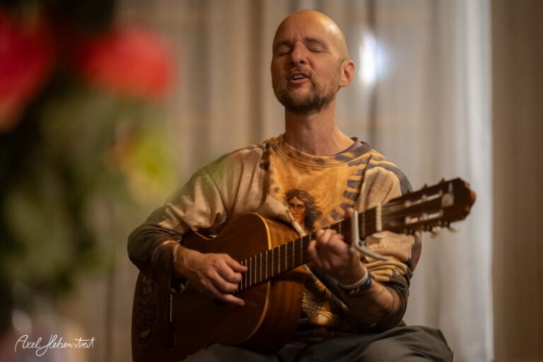 Ein bärtiger Musiker spielt konzentriert Akustikgitarre in warmem Licht vor heller Wand.