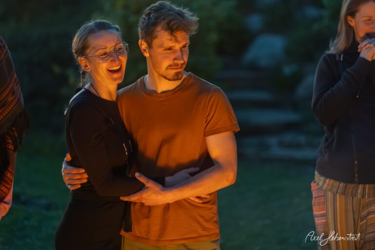 Paar umarmt sich lachend bei goldenem Abendlicht im Freien, umgeben von Freunden bei entspannter Zusammenkunft.