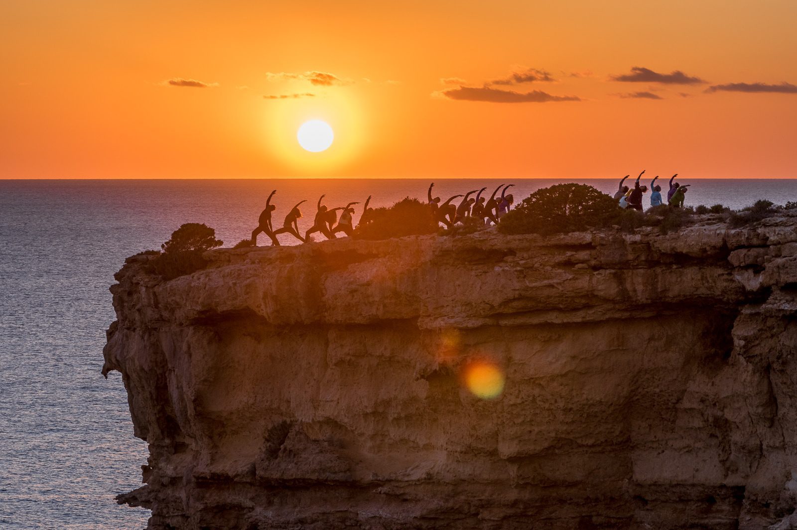 Eine Gruppe von Menschen springt bei Sonnenuntergang silhouettiert auf einer Felsklippe über dem Meer – © Axel Hebenstreit