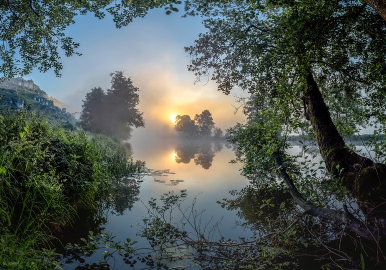 Nebeliger Sonnenaufgang über einem ruhigen See mit Uferbäumen und sanftem Lichtreflex in bayerischer Naturlandschaft – © Axel Hebenstreit