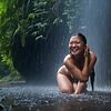 Lächelnde Frau kniet im Regen unter einem Wasserfall im grünen Wald und genießt die erfrischende Naturerfahrung – © Axel Hebenstreit