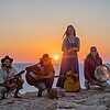 Vier Musikerinnen und Musiker mit Gitarren und Rahmentrommel performen bei Sonnenuntergang am Strand mit glühender Sonne im Hintergrund – © Axel Hebenstreit