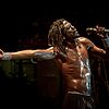 Shirtloser Sänger mit Dreadlocks performt mit ausgestrecktem Arm und Mikrofon auf dunkler Konzertbühne – © Axel Hebenstreit