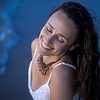 Frau mit geschlossenen Augen lächelt sanft vor blauem Hintergrund, trägt weißes Top und Kettenhalskette – © Axel Hebenstreit