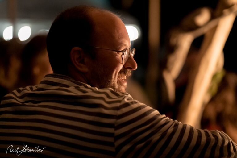 Ein lächelnder Mann mit Brille in einem gestreiften Shirt genießt die Atmosphäre eines abendlichen Events – © Axel Hebenstreit