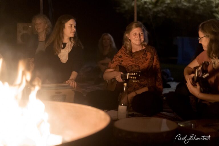 Gemütliche Atmosphäre am Lagerfeuer mit Frauen, die musizieren und lachen, umgeben von Natur – © Axel Hebenstreit