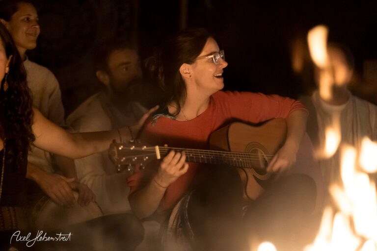 Eine lächelnde Frau spielt Gitarre am Lagerfeuer, umgeben von Zuhörern in gemütlicher Atmosphäre. – © Axel Hebenstreit