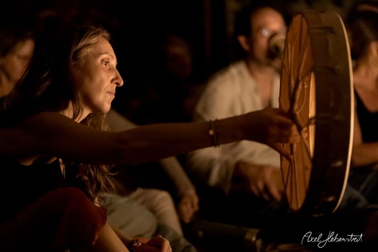 Eine Frau spielt bei schwachem Licht ein Trommelinstrument in einer zusammenhängenden Gruppe. – © Axel Hebenstreit