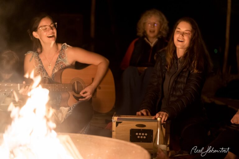 Zwei Frauen singen am Lagerfeuer, während eine Gitarre gespielt wird; eine gemütliche, entspannte Abendatmosphäre. – © Axel Hebenstreit