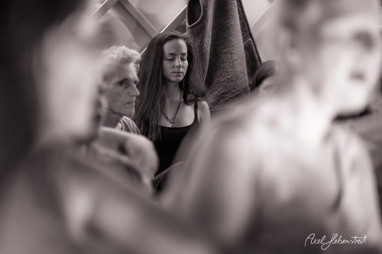 Eine nachdenkliche junge Frau sitzt zwischen anderen Personen und strahlt eine ruhige Stimmung aus. – © Axel Hebenstreit