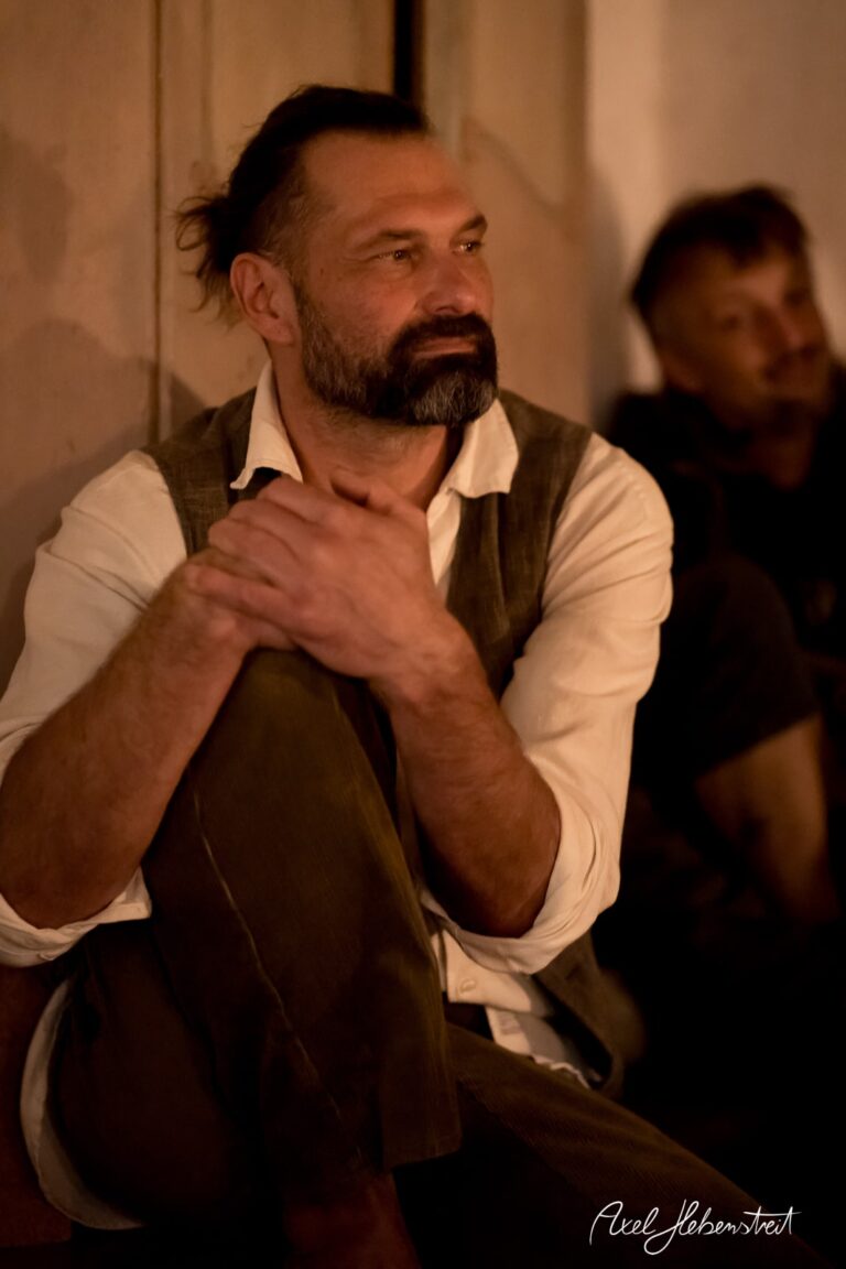 Ein nachdenklicher Mann sitzt mit verschränkten Armen und lächelndem Blick in einem gemütlichen, dimmigen Raum – © Axel Hebenstreit