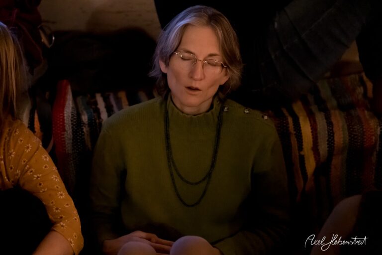 Eine meditierende Frau in einem grünen Pullover sitzt in einer entspannten Atmosphäre auf einem bunten Teppich. – © Axel Hebenstreit