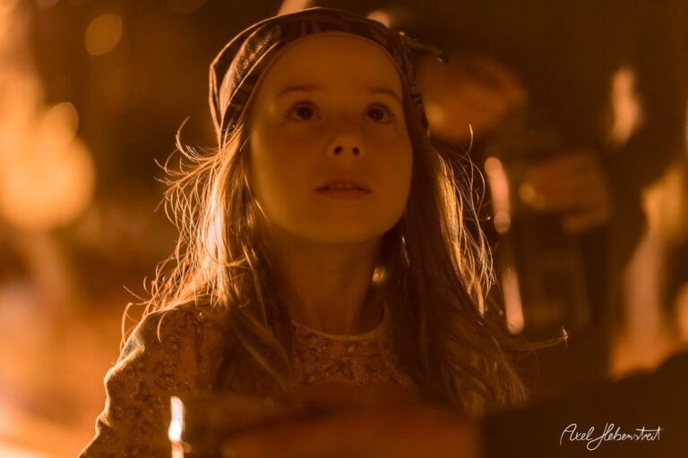Ein Mädchen mit leuchtenden Augen in warmem Licht, neugierig blickend, umgeben von festlicher Stimmung. – © Axel Hebenstreit